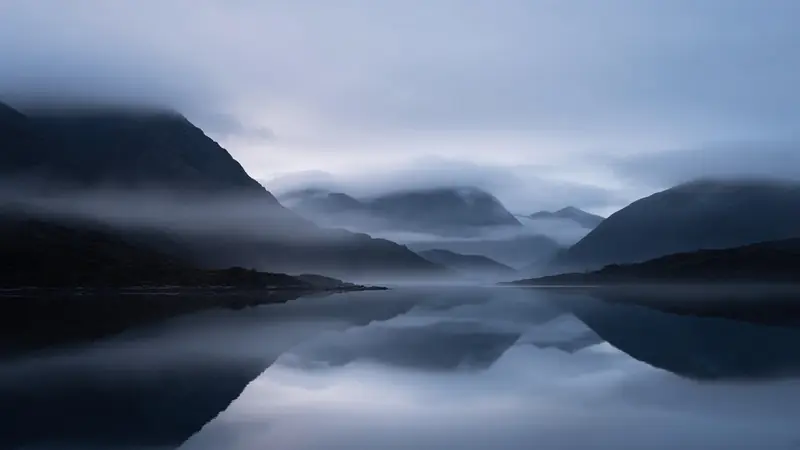 Fog settling over a still fjord at blue hour, dark mountains reflected in the water