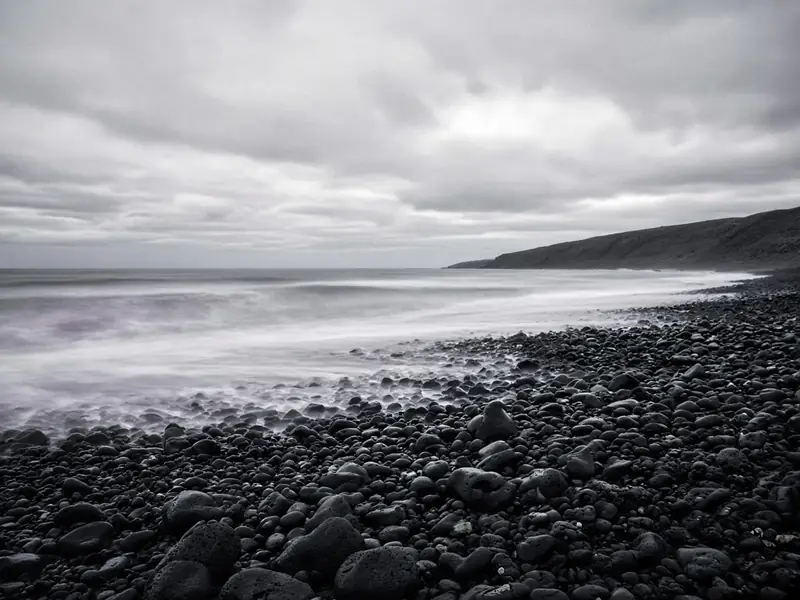 Black basalt pebbles on a beach with soft white surf under an overcast sky