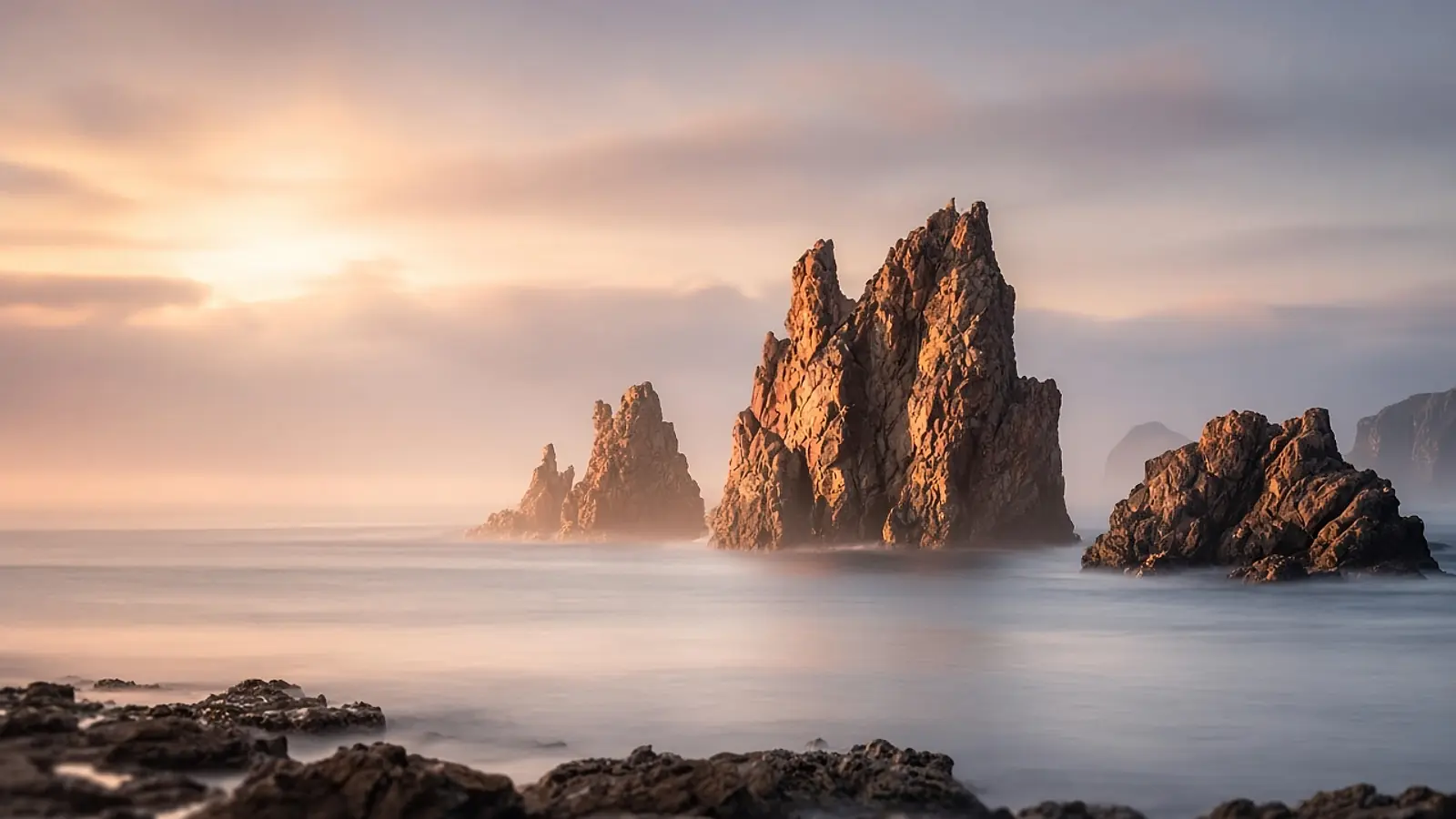 Tall sea stacks lit warm by sunrise above smooth, misty water