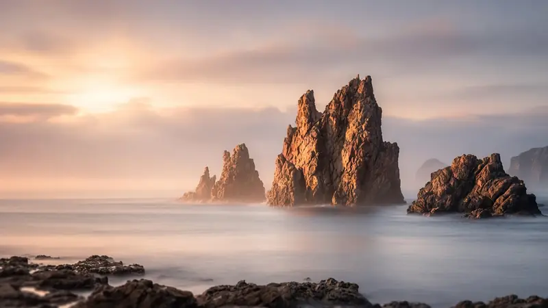 Tall sea stacks lit warm by sunrise above smooth, misty water
